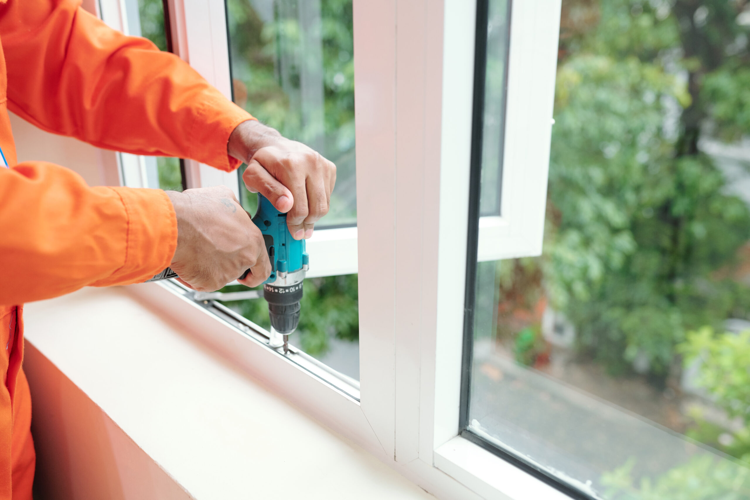 Close-up of unrecognizable technician in orange wear driving screws into jamb of plastic window while installing it
